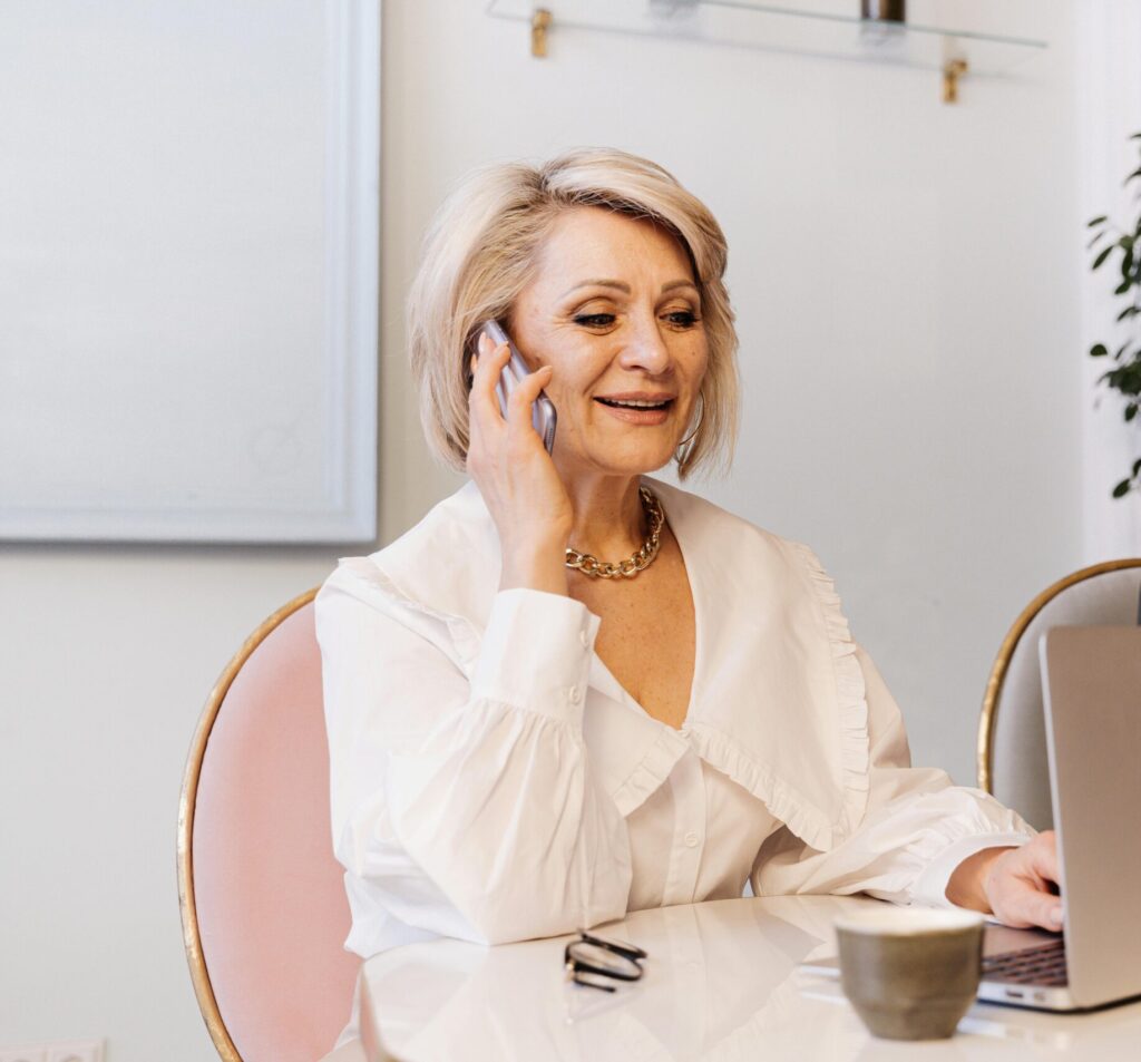 Two senior businesswomen engaged in work, using a phone and a laptop in a modern office.